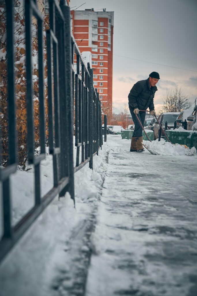 A man wearing winter clothing shovels snow from a sidewalk by a metal fence in front of a building.